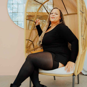 Plus Size Model trägt blickdichte schwarze Overknee-Strümpfe und sitzt mit überschlagenden Beinen in einem Korbsessel