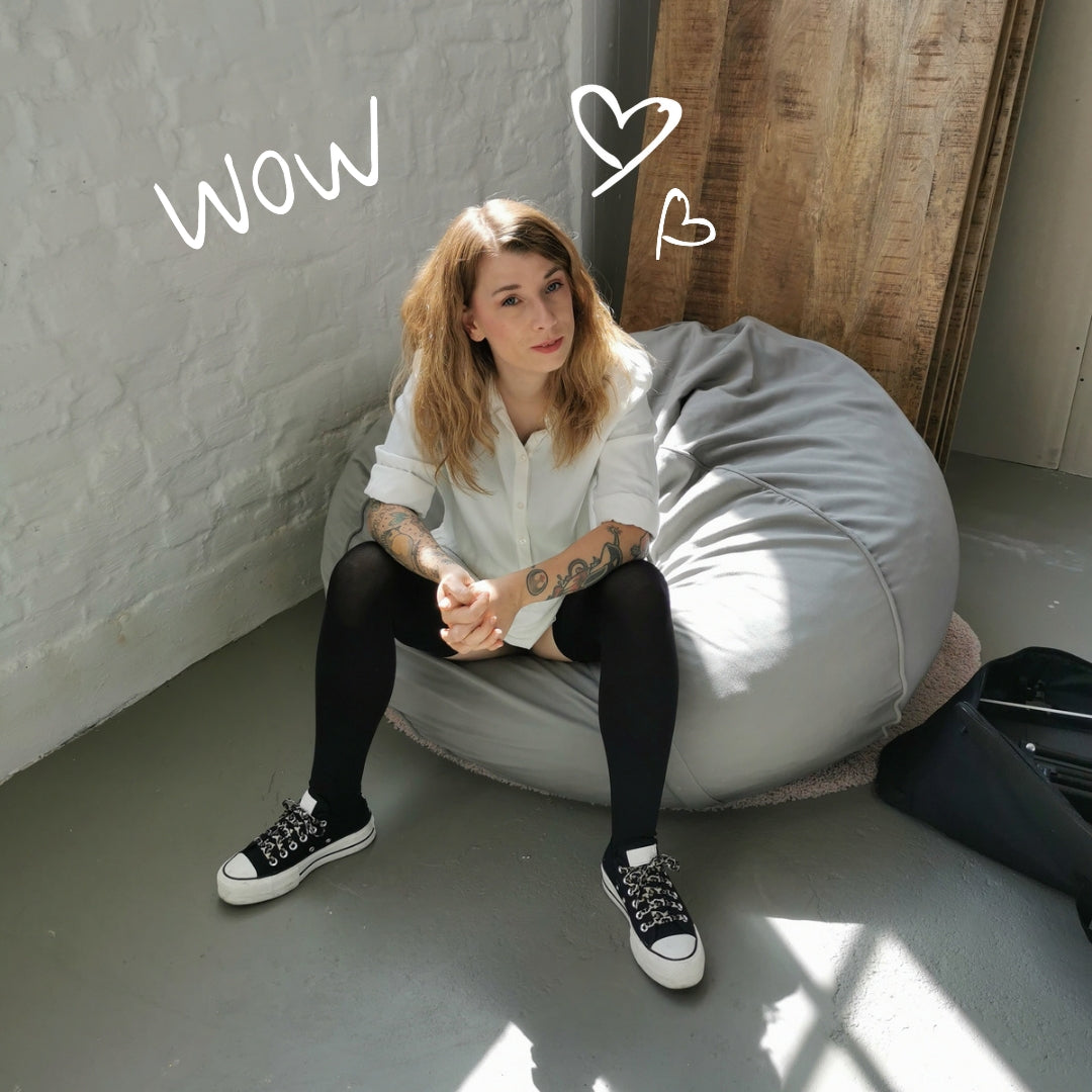 Zierliche, schlanke Frau mit mittelblondem Haar sitzt auf einem grauen Sitzsack. Ihr Outfit besteht aus einem weißen Hemd und schwarzen Overknee-Strümpfen "Defiance". Ihre Schuhe sind schwarz-weiße Sneaker. Sie hat mittelblondes, langes Haar und an ihren Armen sieht man Tattoos. 
