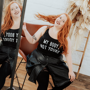 Das Model sitzt auf einem Stuhl vor einem Spiegel. Sie hat rote Haare, roten Lippenstift, trägt ein Top mit der Aufschrift "My Body, My Rules", einen schwarzen Lederrock und Making Waves Strumpfhosen. Lachend wirft sie ihre langen Haare über ihre Schulter.