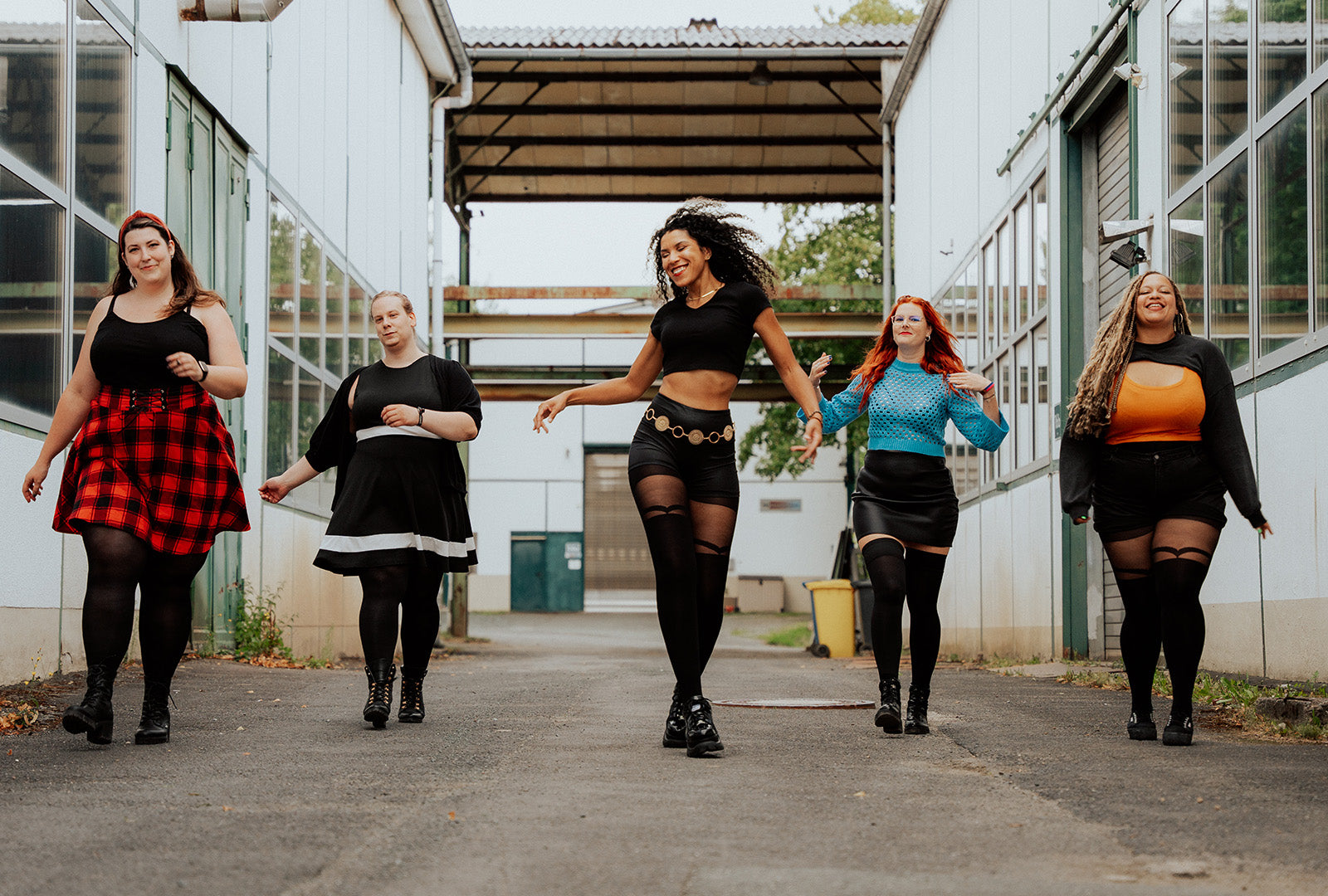 Overknee-Strümpfe und Strumpfhosen an 5 Frauen, die nebeneinander versetzt auf einer Straße zwischen Industriegebäuden laufen.