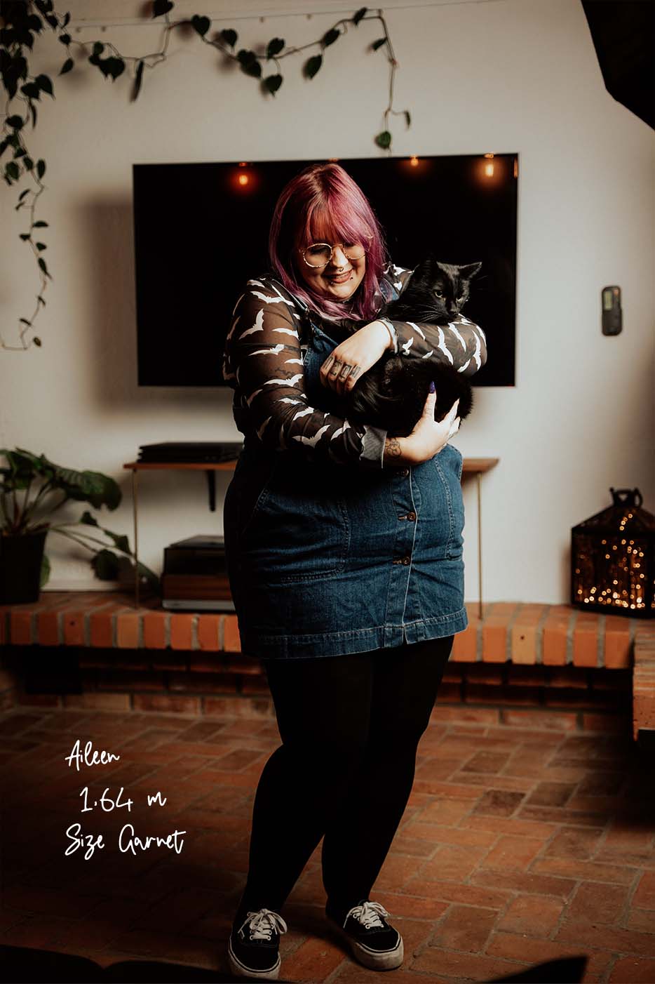 Alltags-Look schwarze Strumpfhose mit Jeanskleid und Mesh-Top an Plus Size Model mit pinken Haaren, die schwarzen Kater hält.