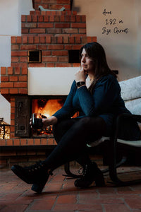 XXL-Strumpfhose blickdicht und schwarz an 182 cm großer Frau, die ein Glas hält. Im Hintergrund ein Kamin mit Feuer.