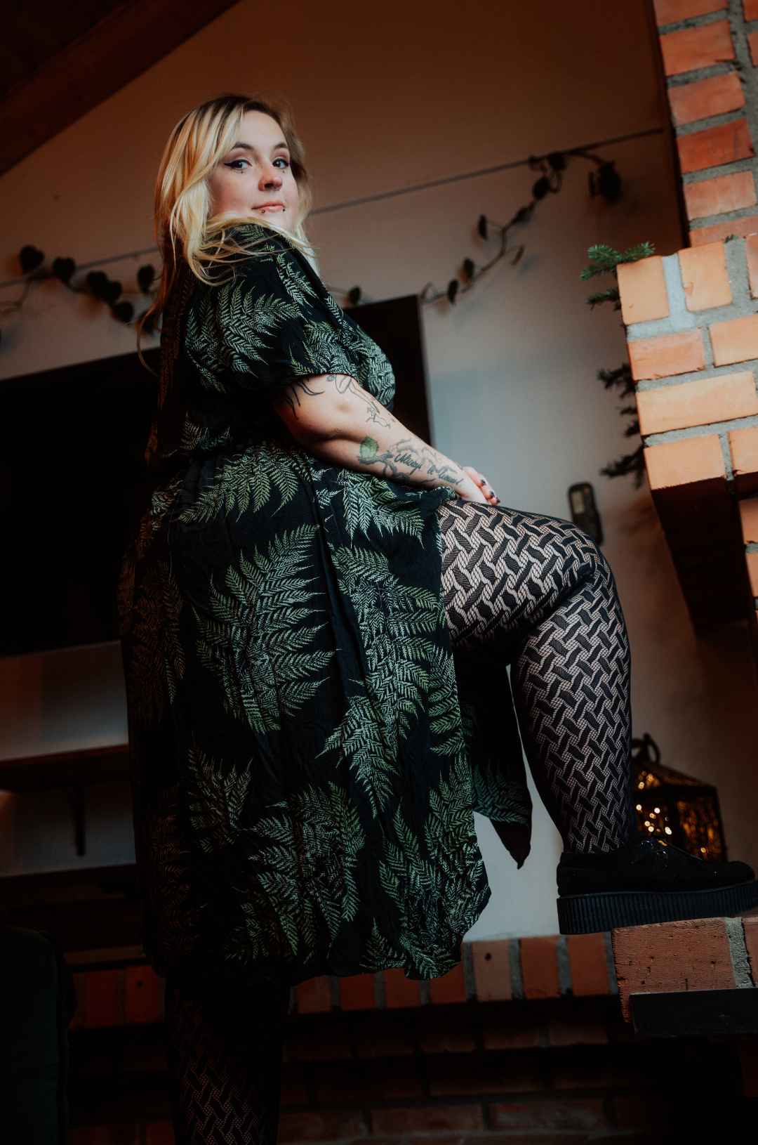 Das blonde Model mit grün-schwarz gemustertem Kleid hat Lipödem, schaut in die Kamera und hat ihr rechtes Bein angewinkelt auf einen Sims gestellt.