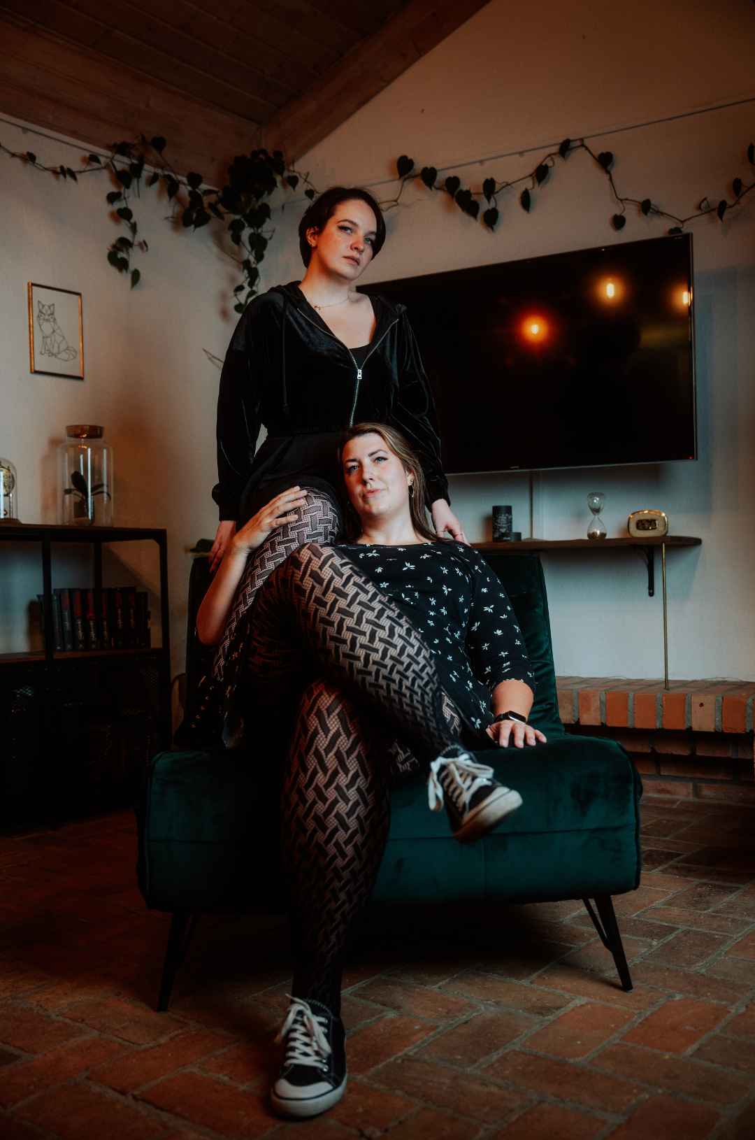 Eine dicke große Frau sitzt auf einem Sessel mit überschlagenen Beinen mit Netzstrumpfhose. Dahinter steht ein zweites Model, das ein Bein - ebenfalls in der Netzstrumpfhose - über die Schulter des sitzenden Models gelegt hat.