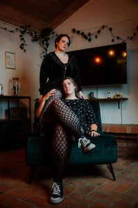 Eine dicke große Frau sitzt auf einem Sessel mit überschlagenen Beinen mit Netzstrumpfhose. Dahinter steht ein zweites Model, das ein Bein - ebenfalls in der Netzstrumpfhose - über die Schulter des sitzenden Models gelegt hat.