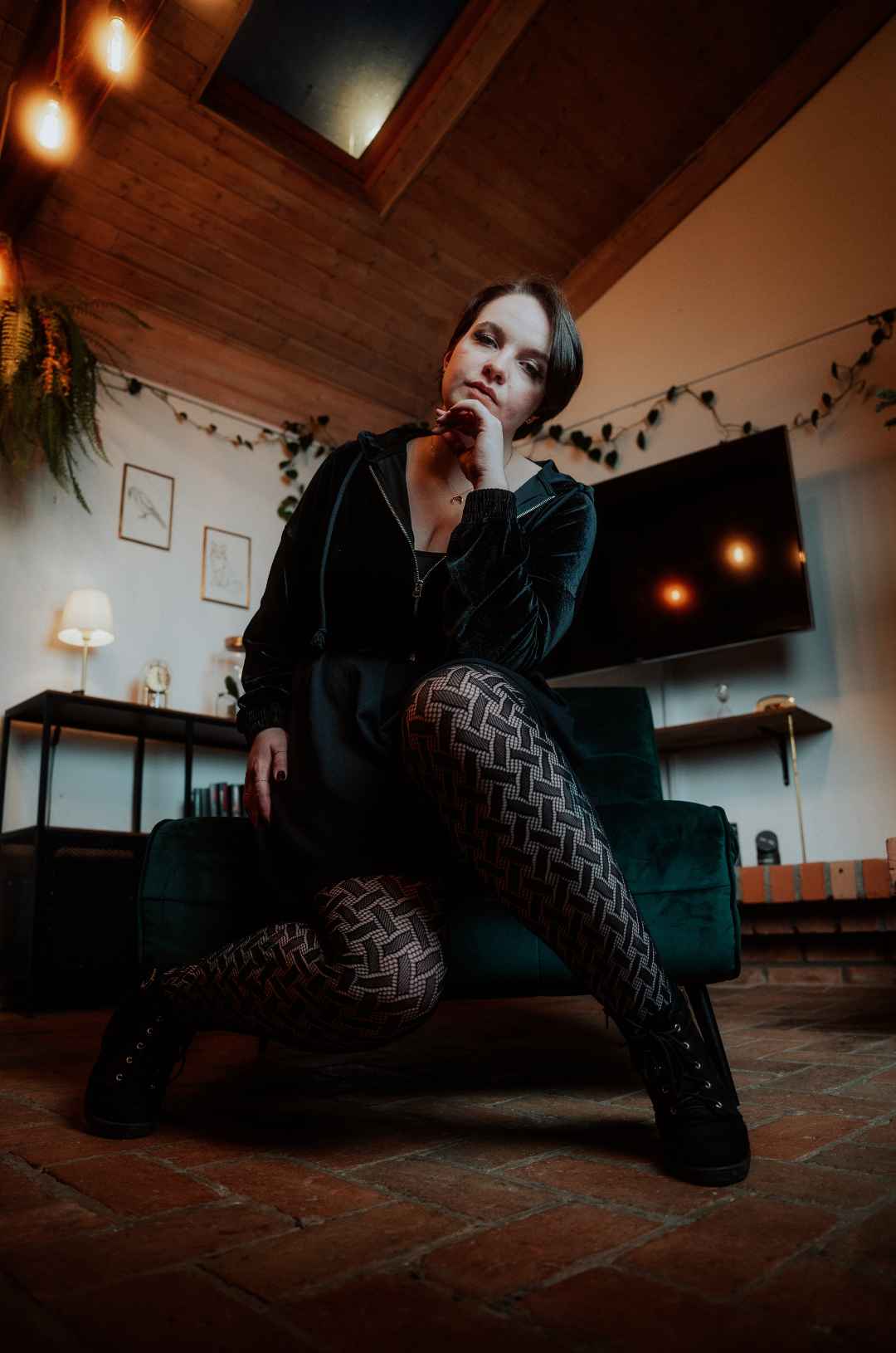 Das Model mit dunklen Haare und dunkler Kleidung schaut von oben herab in die Kamera auf einem Sessel sitzend. Sie trägt eine schwarze Netzstrumpfhose mit Webmuster.