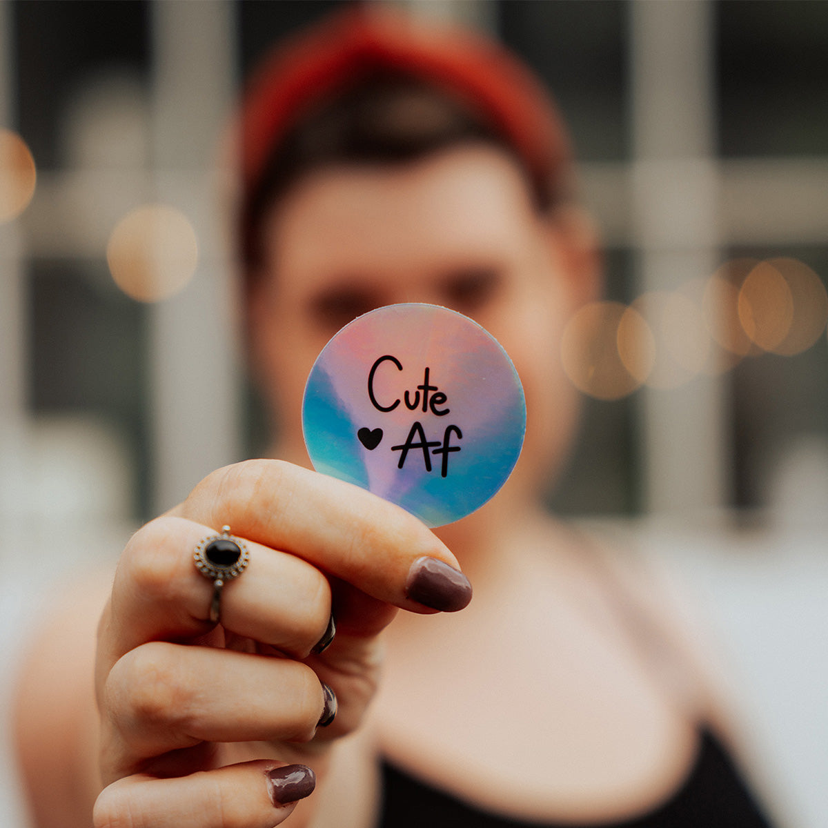 In der Mitte ein runder Holografischer Sticker auf dem in schwarzer Schrift "Cute Af" mit einem kleinen Herzchen zu sehen ist. Der Sticker wird von einer Hand gehalten mit einem Ring am Mittelfinger und braunem Nagellack. Im Hintergrund sieht man verschwommen eine Frau mit rotem Haarreifen und schwarzem Spaghetti-Top. Dahinter eine Lichterkette und weiße Fensterstreben. 