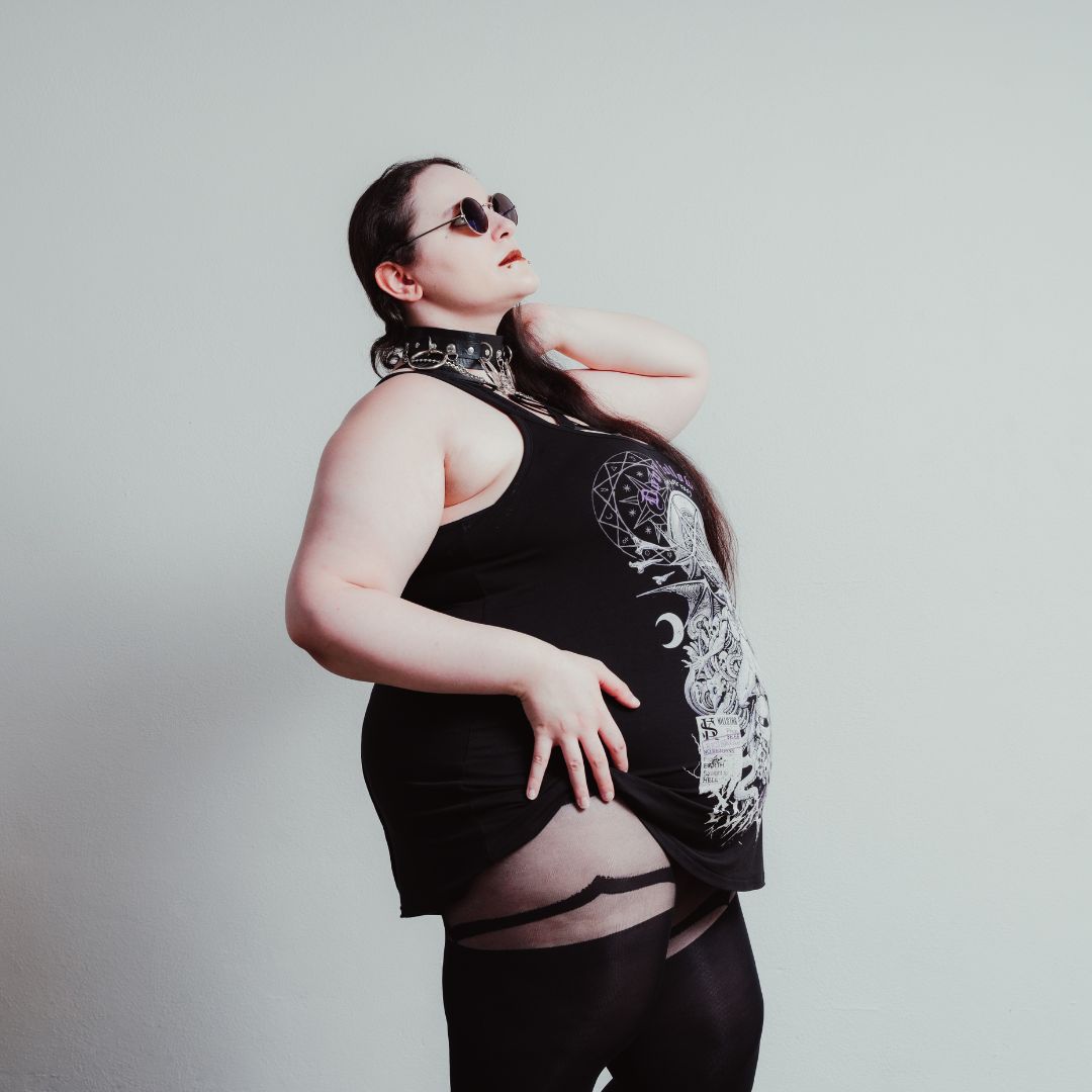 Eine Frau steht in Strumpfhosen und mit schwarzem Top nach rechts gerichtet und hat ihren Kopf nach oben gerichtet. Sie trägt eine schwarze Sonnenbrille mit runden Gläsern.